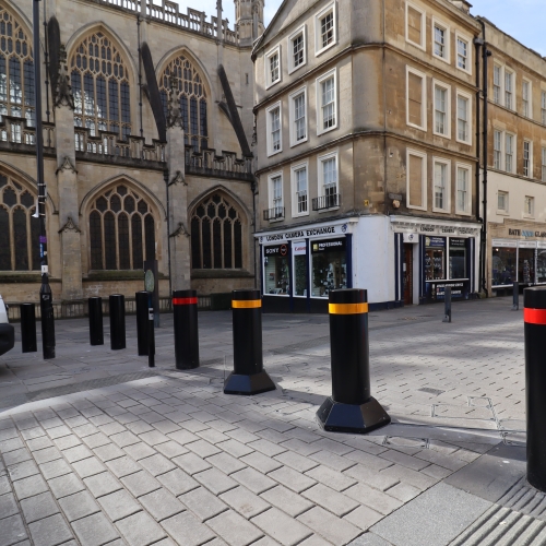 Black bollards with red and yellow bands on a cobbled street, in front of a historic building with Gothic windows and nearby shops